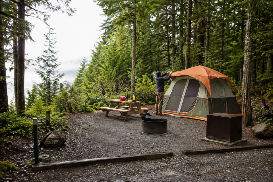 A camper setting up an orange tent in the forest beside a fire pit