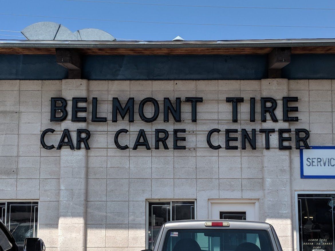 Belmont Tire Car Care Center sign on a gray building with parked vehicles below