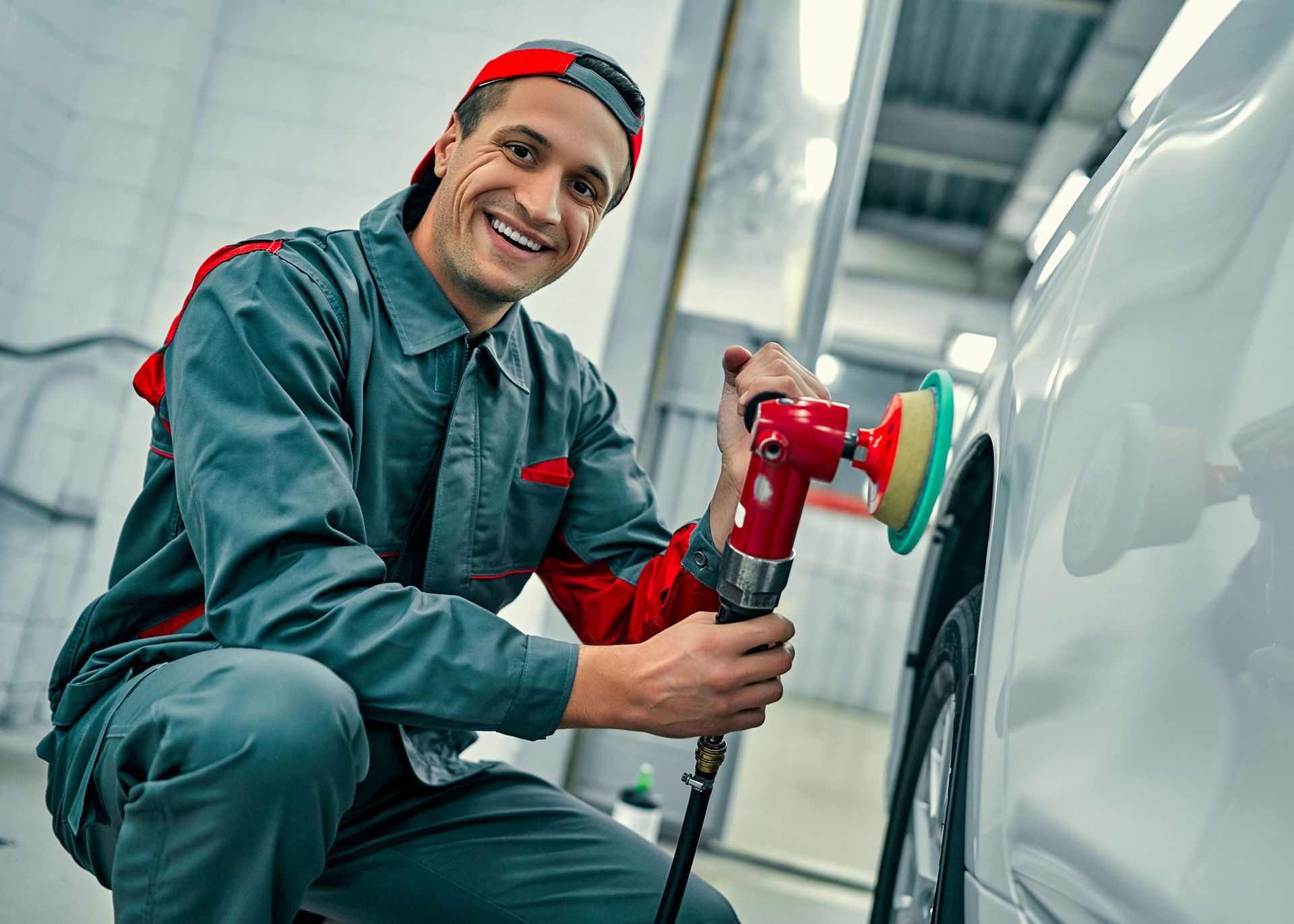 Smiling mechanic polishing car's side panel in a garage. He's wearing blue overalls and a red cap.