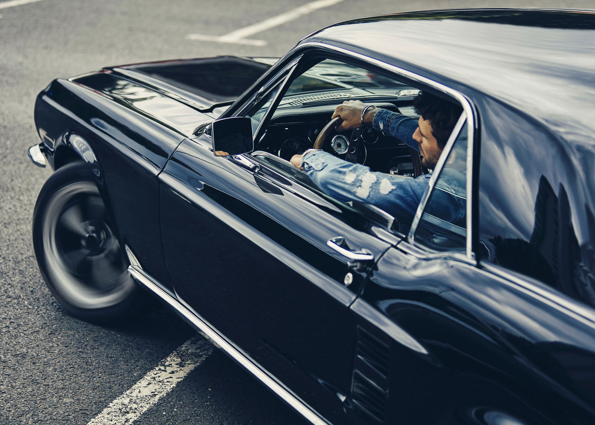 Man driving a classic black car, parked on a paved surface.