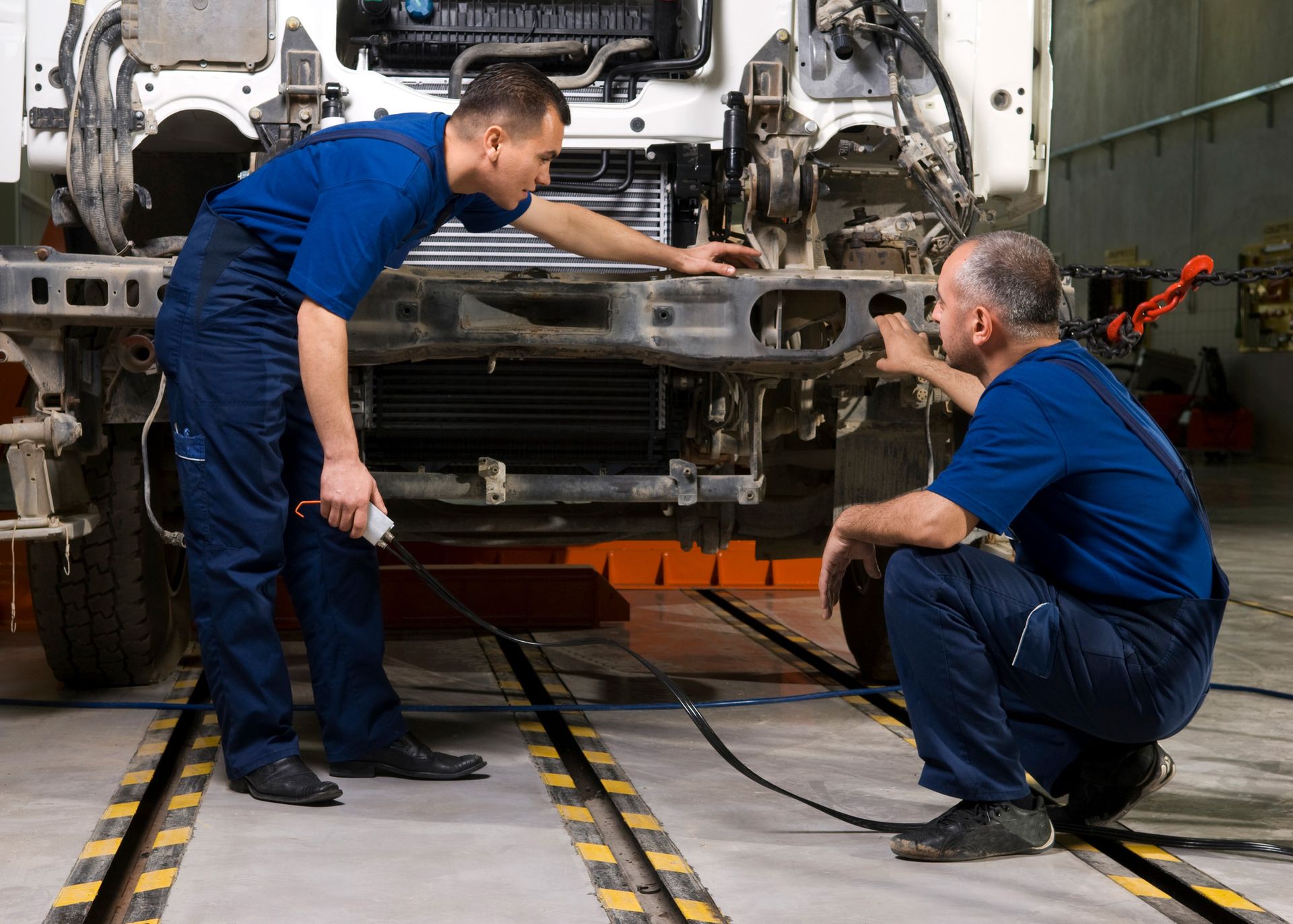 Two mechanics in blue discuss a truck's front end in a repair shop. One stands, pointing; the other squats.
