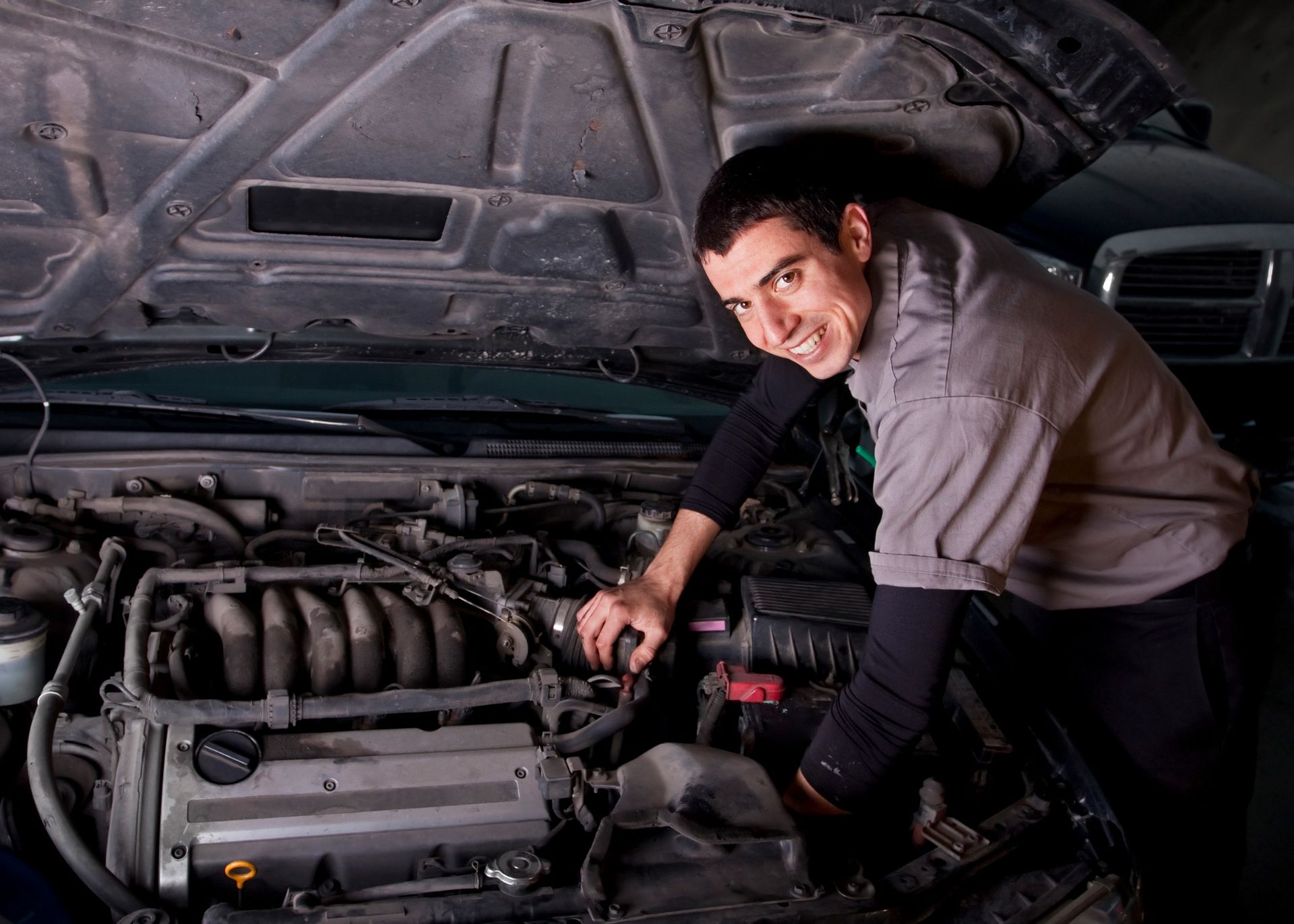 Mechanic smiling while working on a car engine with the hood open.