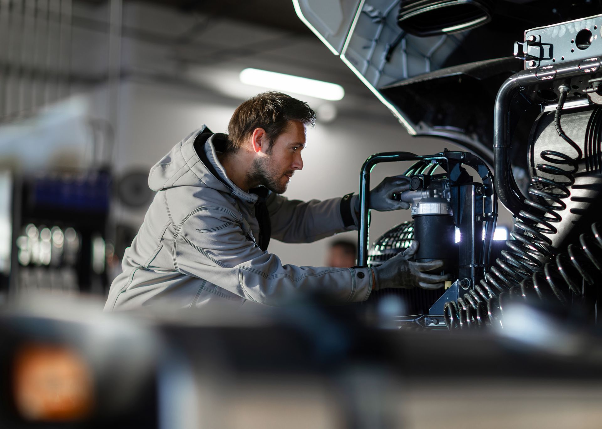 Mechanic working on a truck engine, wearing a gray jacket, in a dimly lit garage.