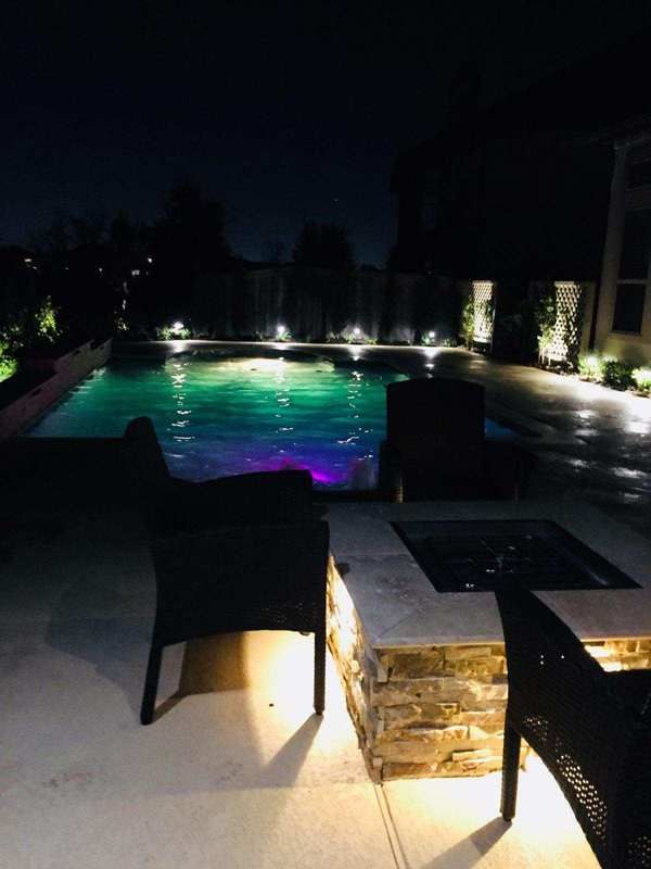 A swimming pool is lit up at night with a fire pit in the foreground.