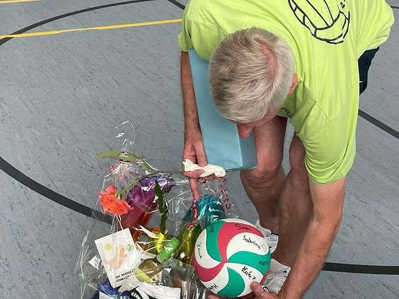 Elmar hält zum Abschied als Trainer einen Volleyball, ein Geschenk und Blumen in einer Turnhalle.