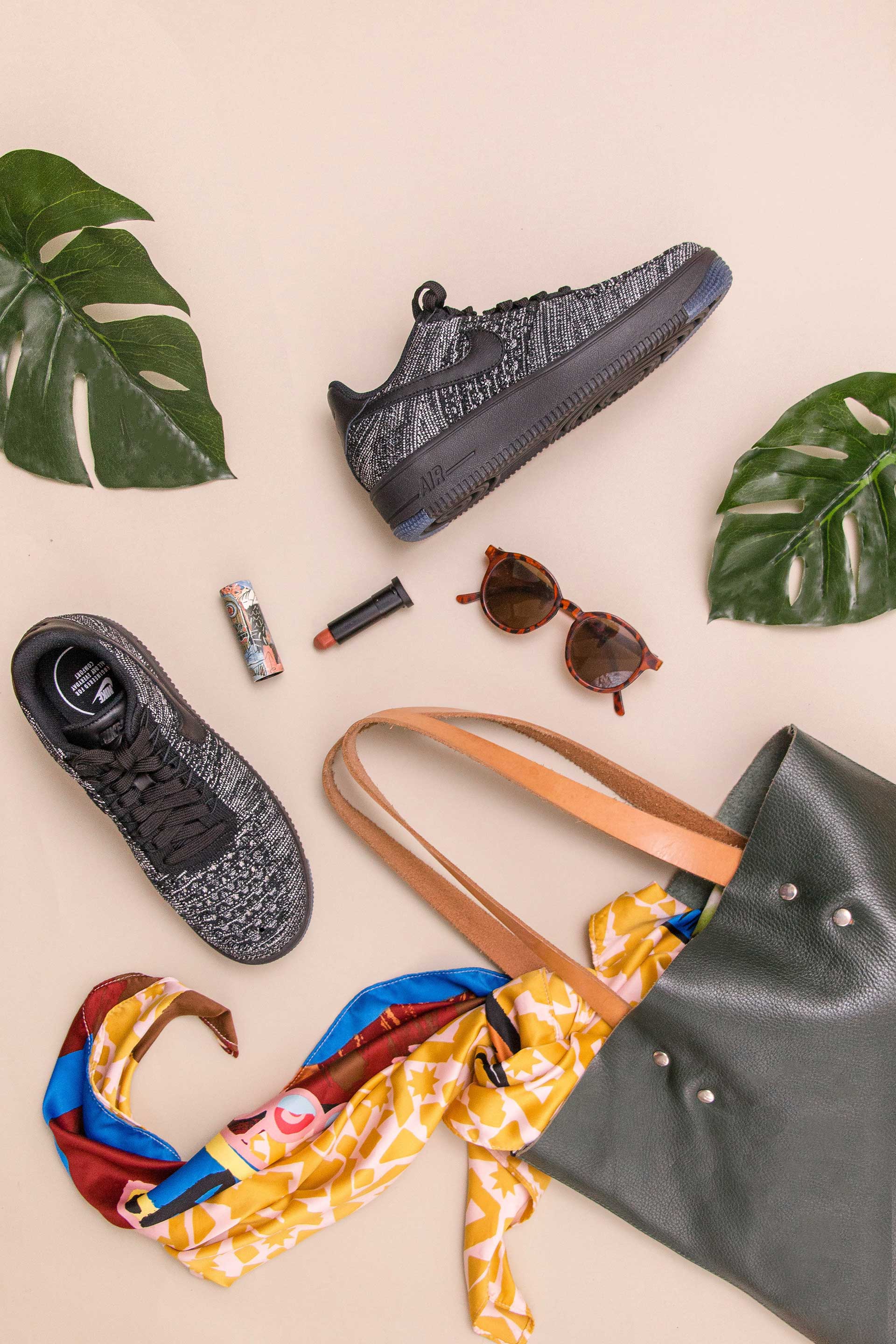 A pair of shoes , a bag , sunglasses , a scarf , and a lipstick are on a table.