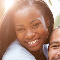 Smiling woman with dark hair, close-up with a man's face on the right, outdoors with sunlight.