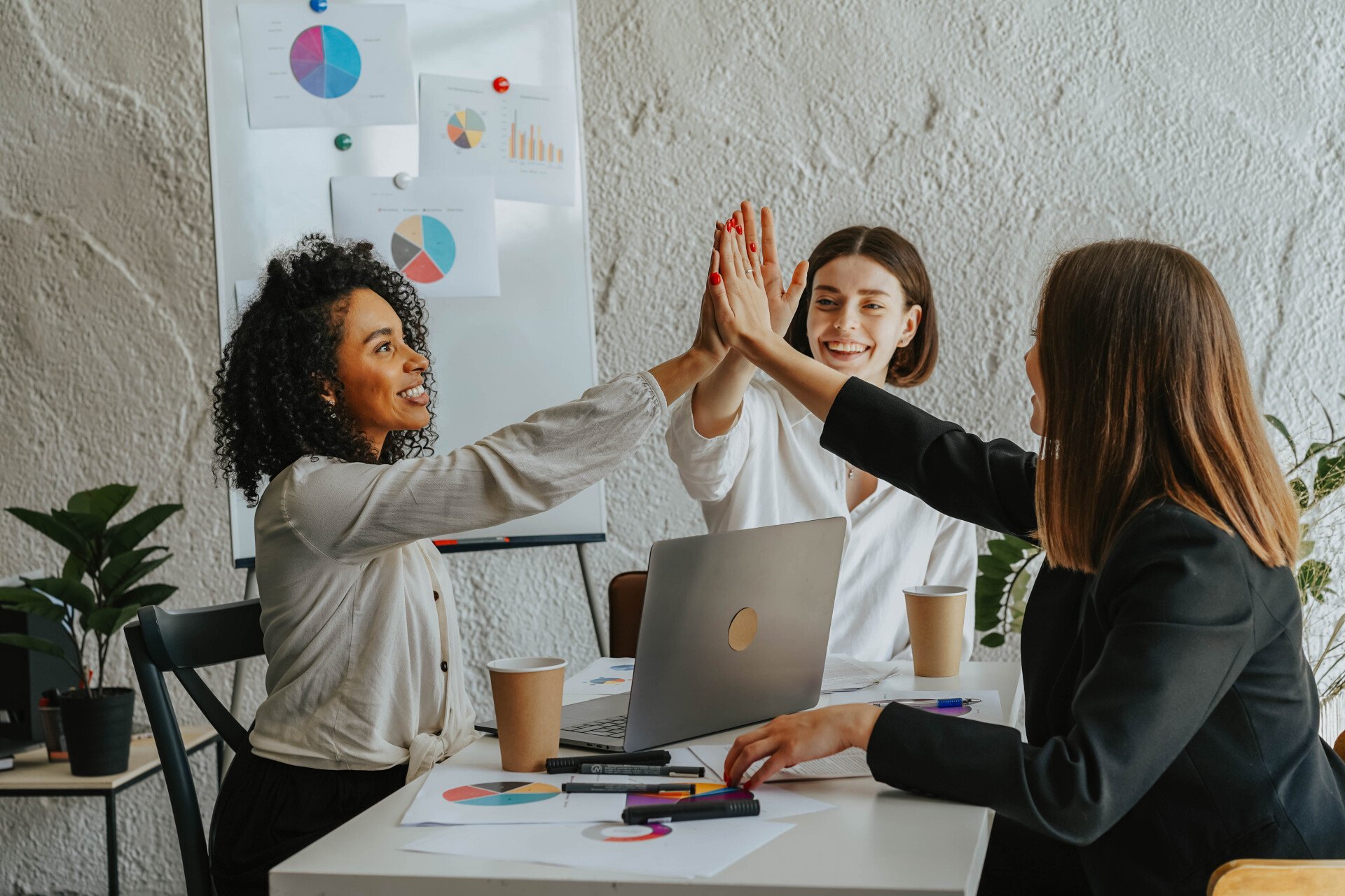 A group of women are giving each other a high five while sitting at a table with laptops.
