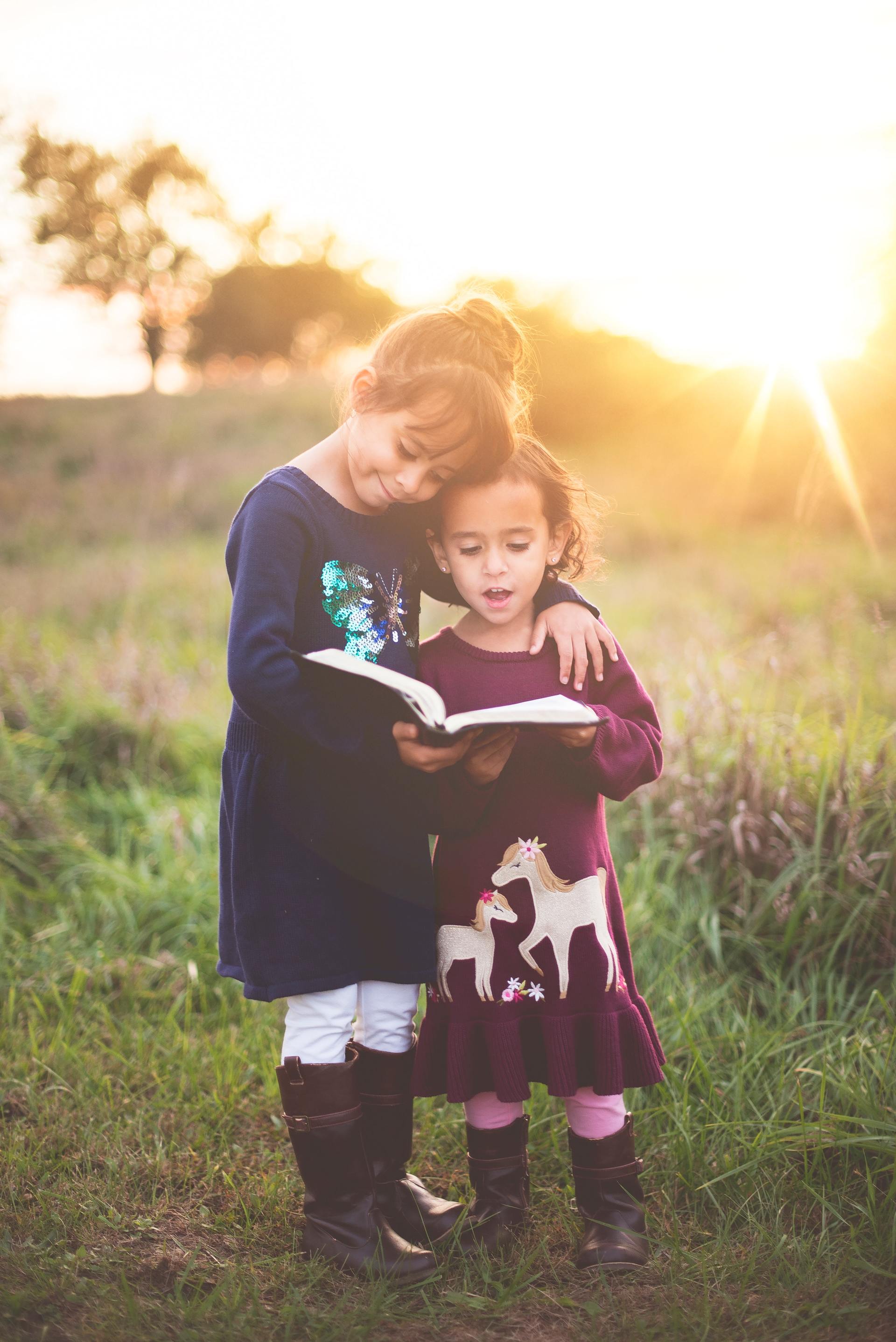 Two young girls reading a book in a field with sunset; one has arm around the other's shoulders.